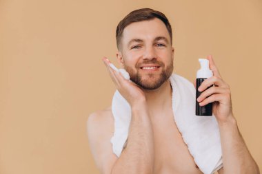 Boynunda havluyla bej arka planda yüz yıkama köpüğü süren sevimli, mutlu sakallı bir adam. Güzellik ve bakım konsepti