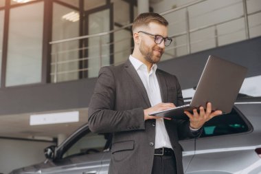 Araba galerisinde dizüstü bilgisayar kullanan gözlüklü ve takım elbiseli modern sakallı bir adam.