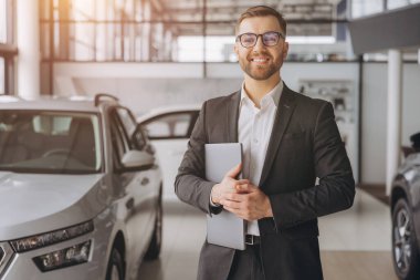 Araba galerisinde dizüstü bilgisayar kullanan gözlüklü ve takım elbiseli modern sakallı bir adam.