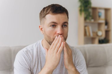 Sakallı, hasta, üzgün bir adam evdeki kanepede oturuyor. Burun nezlesi salgını, koronavirüs salgını, çoban salgını, hapşırma....