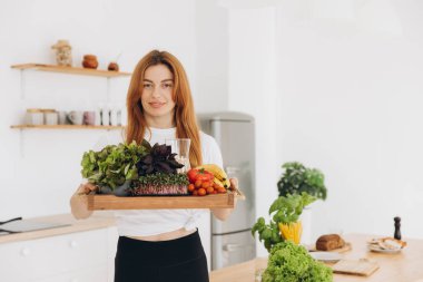 Happy sporty mature woman smiling and holding tray with healthy food in kitchen at home