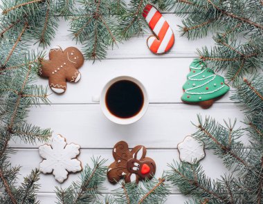 Beyaz ahşap bir masada bir fincan kahve setiyle zencefilli adam ve ağaç şeklinde Noel kurabiyeleri. Etrafı yemyeşil dallarla çevrili..
