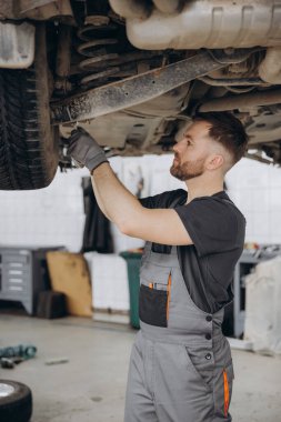 Yoğunlaştırılmış araba servisi çalışanının otomobil kontrol ekipmanlarına bakın. Üniformalı ve eldivenli erkek tamirci arabanın altında duruyor ve kırılma noktası arıyor.