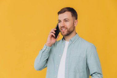 Sarı arka planda izole edilmiş, günlük mavi gömlekli neşeli genç adam, stüdyo portresi. İnsanlar samimi duygular, yaşam tarzı konsepti. Kopyalama alanı oluşturun. Cep telefonuyla konuşuyor.