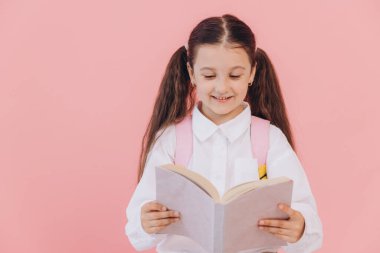 Sırt çantasıyla pembe arka planda kitap okuyan, okul üniformalı tatlı, güler yüzlü küçük kız.