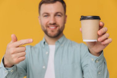 Nane rengi gömlekli, elinde kahve fincanı olan ve sarı arka plandaki modelin için fotokopi alanını gösteren tıraşsız yakışıklı bir adam.