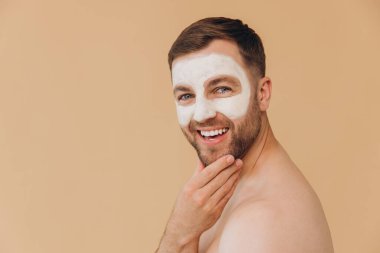 Tıraşsız, gülümseyen bir adamın güzel portresi bej renkli stüdyo arka planına yüz bakımı maskesi takıyor. Dermatoloji, lüks spa prosedürleri. Erkek güzellik anlayışı, kozmetik ürünler..