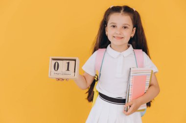 Birinci sınıftaki küçük bir kızın portresi kameraya bakıyor ve 1 Eylül 'de izole edilmiş sarı arka planda ahşap bir takvim tutuyor. Okula geri dön.