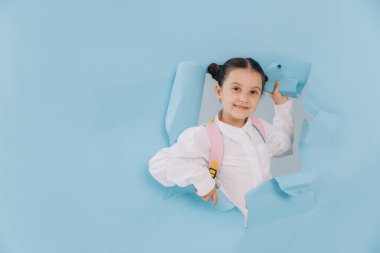 Güzel küçük kız yırtık kağıt delikten mavi bir stüdyonun arka planına bakıyor.