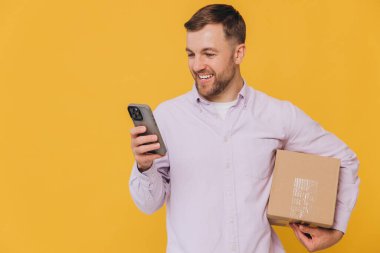 Sıradan kıyafetli, portresiz yakışıklı bir adam, elinde bir kutu ve mutlu bir şekilde akıllı telefondan online alışveriş yapıyor, izole edilmiş sarı arka planda heyecanlı bir gülümsemesi var.