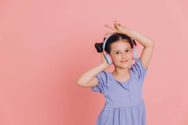 Mavi elbiseli sevimli beyaz çocuk müzik dinliyor kablosuz kulaklık takıyor ve dans ediyor, pembe arka planda izole edilmiş.