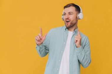 Tıraşsız, temiz gömlekli, kulaklıkla müzik dinleyen ve sarı arka plandaki fotokopi alanını gösteren yakışıklı bir adam.