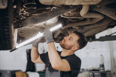 Yoğunlaştırılmış araba servisi çalışanının otomobil kontrol ekipmanlarına bakın. Üniformalı ve eldivenli erkek tamirci arabanın altında duruyor, lambayı tutuyor ve kırılma noktası arıyor.