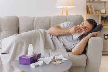 Evdeki kanepede sıcak bir battaniyenin altında yatıp kağıt mendille hapşıran hasta, beyaz bir adam. Soğuk algınlığı, grip, mevsimsel kış enfeksiyonları