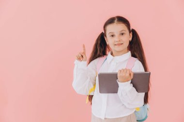 Sırt çantalı, tabletli ve pembe arka planda parmağı olan sevimli, gülümseyen küçük kız.