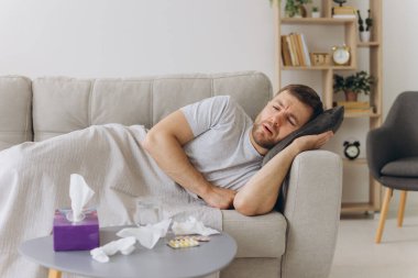 Kafkasyalı hasta, başı ağrıyan bir adam sıcak bir battaniyenin altında evdeki kanepede ellerini acı içinde tutuyor. Soğuk algınlığı, grip, mevsimsel kış enfeksiyonları