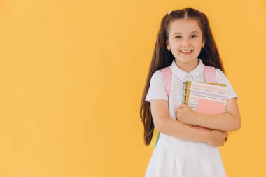 Sırt çantalı, sarı arka planda defterleri ve kitapları olan beyaz okul üniformalı küçük tatlı kız.