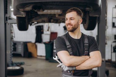 Kollarını kavuşturup araba servisinde İngiliz anahtarı tutarken gülümseyen sakallı bir tamirci.