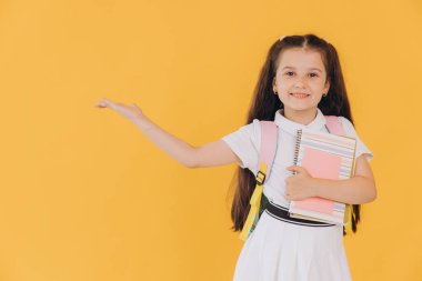 Beyaz okul üniformalı, elinde not defterleri olan ve sarı arka planda eli olan küçük tatlı bir kız.