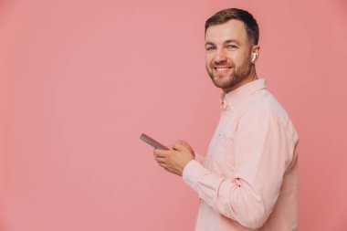 Yan görünüm mutlu beyaz sakallı adam pembe gömlek giyer günlük kıyafetler giyer kulaklıkla müzik dinler basit pastel açık pembe arka plan stüdyo portresi üzerinde izole edilmiş telefona bakar