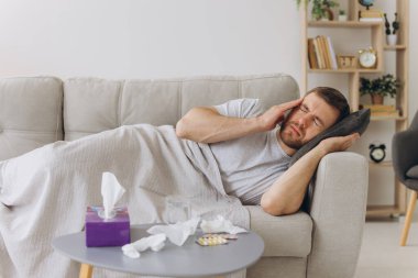 Kafkasyalı hasta, başı ağrıyan bir adam sıcak bir battaniyenin altında evdeki kanepede ellerini acı içinde tutuyor. Soğuk algınlığı, grip, mevsimsel kış enfeksiyonları