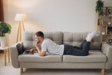 Bir beyaz erkek, evde kulaklıkla bilgisayar başında çalışıyor. Müşteri hizmetleri müdürü evde kanepede gömlek ve pijama giyerek yatıyor.