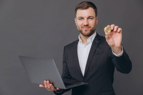 Portrait of a cheerful businessman showing bitcoin while holding laptop computer isolated over gray background