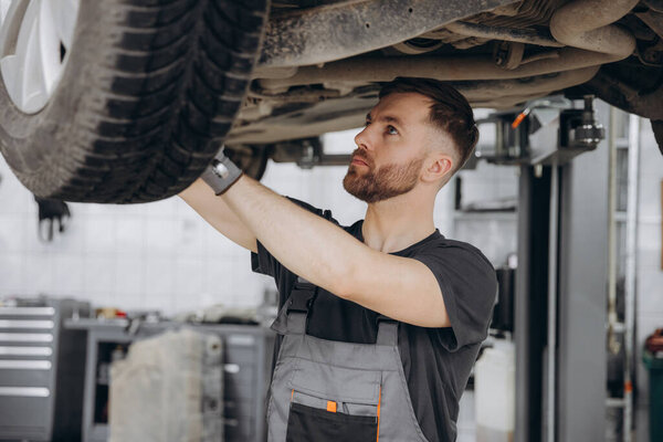 Вид сбоку концентрированного работника автосервиса, проверяющего ходовой механизм автомобиля. Мужчина-механик в форме и перчатках стоит под машиной и ищет поломки
