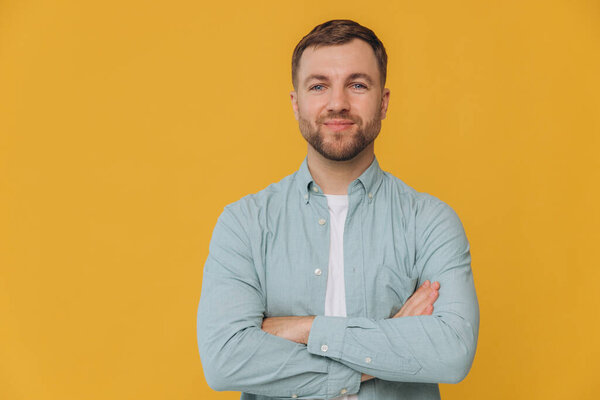 Cute unshaven man smiling with healthy teeth and crossed arms in mint shirt posing isolated on yellow background with copy space