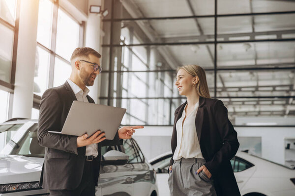 Продавец-мужчина показывает что-то на ноутбуке клиентке, покупающей машину в автосалоне.