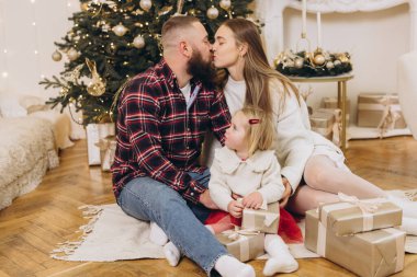 Anne babalar Noel ağacının altında kızlarıyla yerde otururken öpüşüyorlar.