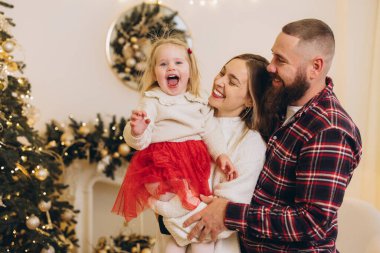 Sakallı anne ve baba, Noel arifesini dekore edilmiş bir evde neşeli kızlarını kucağında tutuyorlar.