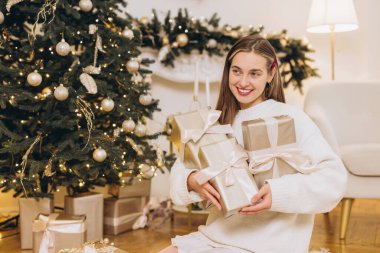 Bir sürü Noel hediyesi taşıyan genç bir kadın, Noel ağacının yanında oturuyor, gülümsüyor ve başka tarafa bakıyor.