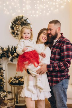 Sakallı baba ve zarif anne sarışın kızlarını kucaklıyor, Noel 'i dekore edilmiş bir evde birlikte kutluyorlar.