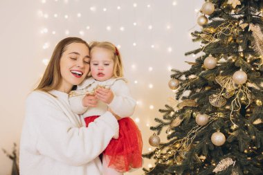 Anne ve kızı gülümsüyor ve Noel ağacını altın süslemeler ve peri ışıklarıyla süslüyorlar, birlikte Noel 'in tadını çıkarıyorlar.