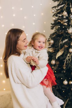 Anne, süslenmiş bir Noel ağacının yanında kırmızı etek giymiş, gülümseyen kızını kucağında tutuyor.