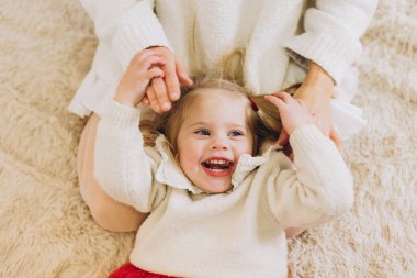 Beyaz süveter ve kırmızı etek giyen küçük bir kız Noel sabahı sıcak bir halıda annesiyle oynarken gülüyor.