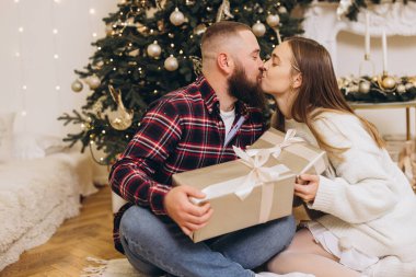 Romantik çift, Noel hediyelerini alırken öpüşüyor. Ev ortamında süslü bir Noel ağacının yanında oturuyor.