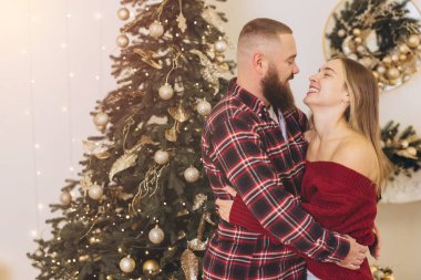 Sakallı adam ve sarışın kadın süslü Noel ağacının yanında sarılıp gülümsüyorlar, birlikte Noel tatilinin tadını çıkarıyorlar.