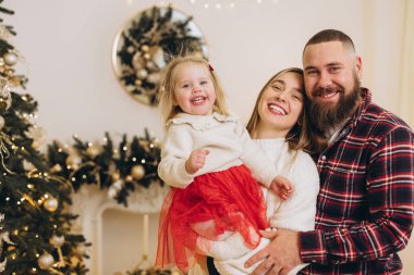 Gülümseyen ebeveynler mutlu kızlarını Noel süslemeli bir oturma odasında tutuyorlar, birlikte neşeli aile zamanlarının tadını çıkarıyorlar.