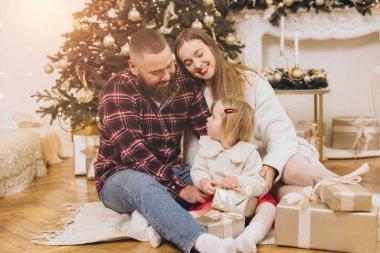 Mutlu bir aile yerde oturuyor ve ev dekore edilmiş bir evde Noel hediyelerini açıyor.