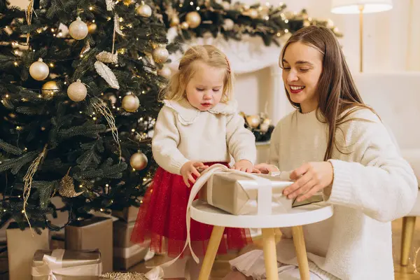 Gülümseyen anne ve kız Noel hediyesini dekore edilmiş ağacın yanında açıyorlar, birlikte bayram sezonunun tadını çıkarıyorlar.