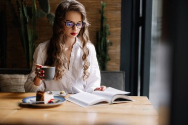 Genç iş kadını bir fincan kahve içip modern bir kafede kitap okuyor.