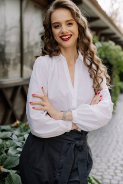 Çapraz kollarla poz veren neşeli bir iş kadınının portresi, beyaz gömlek ve siyah pantolon giyiyor, özgüven ve profesyonellik sergiliyor.