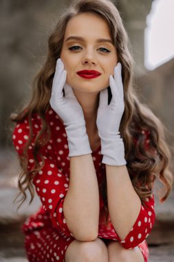 Kırmızı rujlu, beyaz eldivenli, kırmızı benekli bir elbise giymiş, yüzüne zarafetle dokunan bir model.
