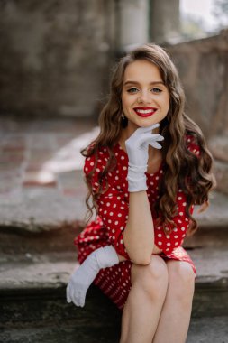 Kırmızı rujlu, gülümseyen, basamaklarda poz veren, klasik kırmızı benekli bir elbise ve beyaz eldivenle güzel bir aktris.