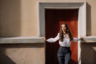 Uzun kahverengi saçlı ve kırmızı rujlu bir iş kadını. Şehirdeki eski bir binanın kırmızı kapısının önünde poz veriyor.