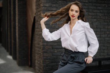 Genç iş kadını bankta oturuyor, saçlarıyla oynuyor, beyaz bir gömlek ve gri pantolon giyiyor, kendine güveni ve tarzı var.