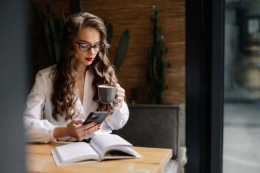 Genç iş kadını kahve içiyor ve kafede uzaktan çalışırken akıllı telefon kullanıyor.