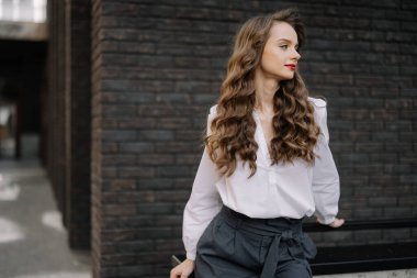 Uzun kahverengi kıvırcık saçlı ve kırmızı rujlu zarif iş kadını şehir ortamında siyah tuğla duvarın yanında poz veriyor.